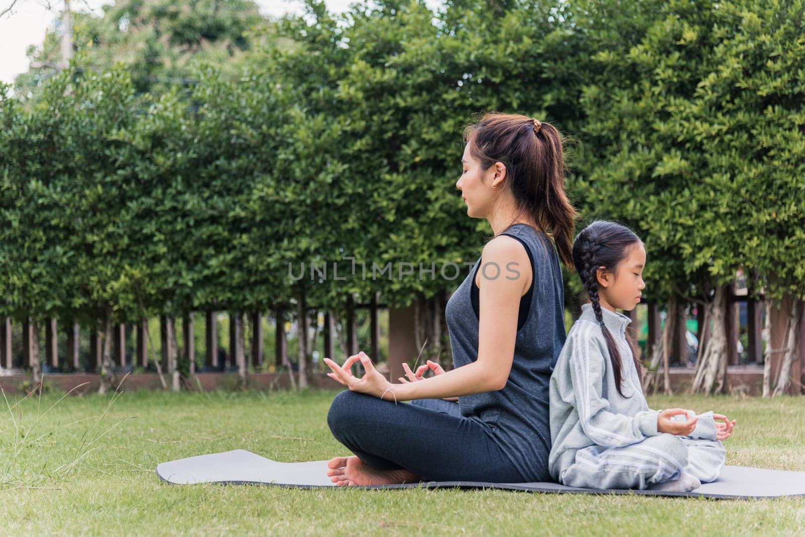 Asian young mother practicing doing yoga exercises with her daughter outdoors in meditate pose together on green grass in nature a field garden park, family sport and exercises for healthy lifestyle