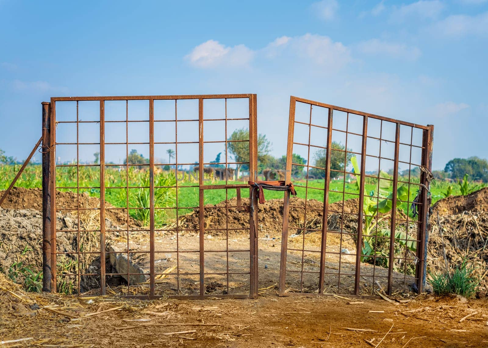 Weathered old gate tied with rope and installed near heaps of soil against cloudy sky on sunny day on farm