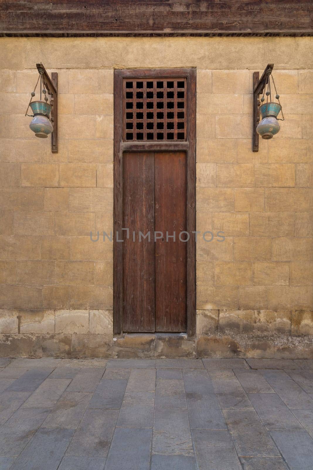 Wooden closed door and two Arabic glass street lanterns hanged on a wooden pole in old stone bricks wall, Medieval Cairo, Egypt
