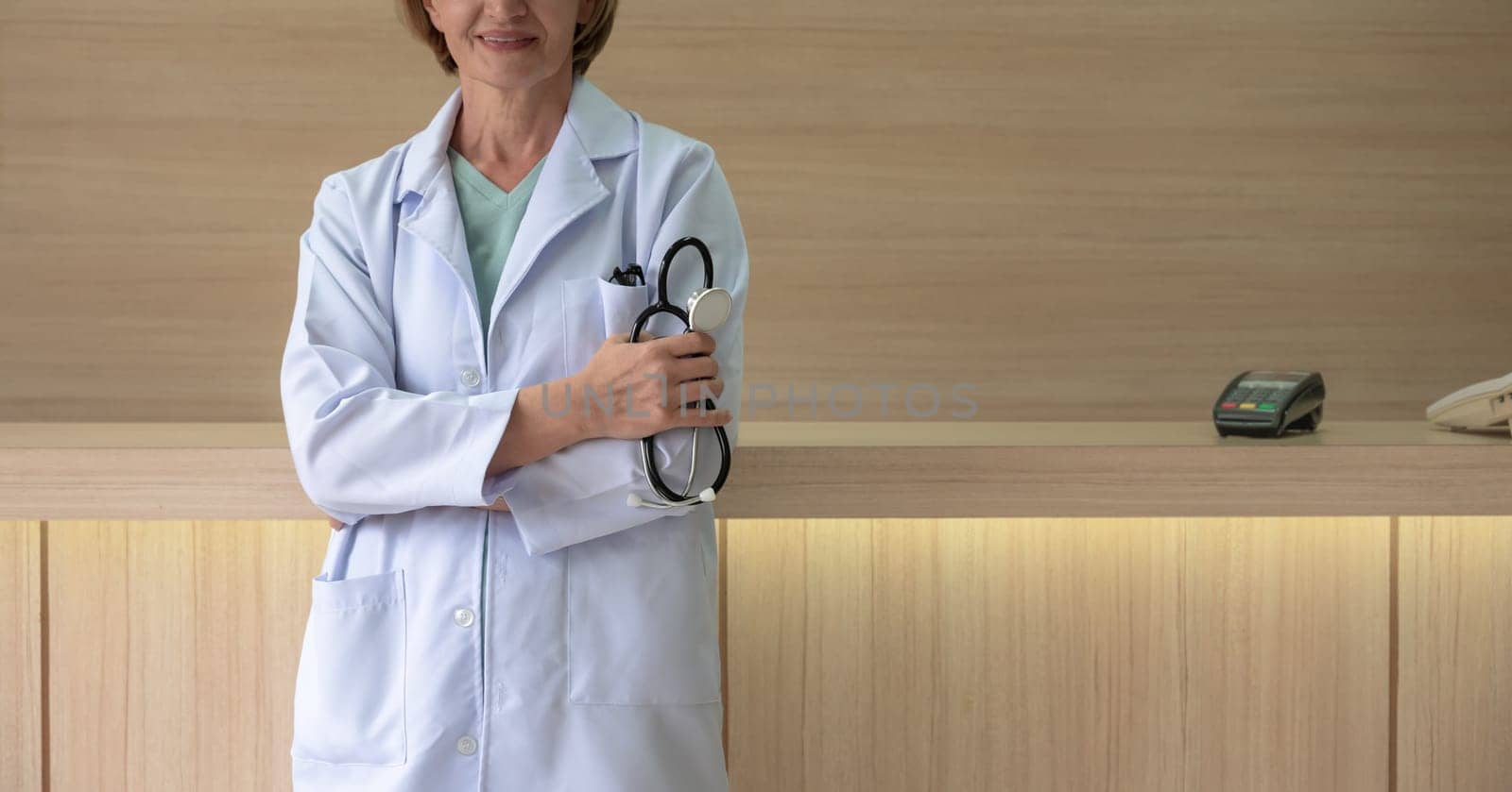 Female doctor in white lab coat holding stethoscope and standing with crossed arms at medical clinic