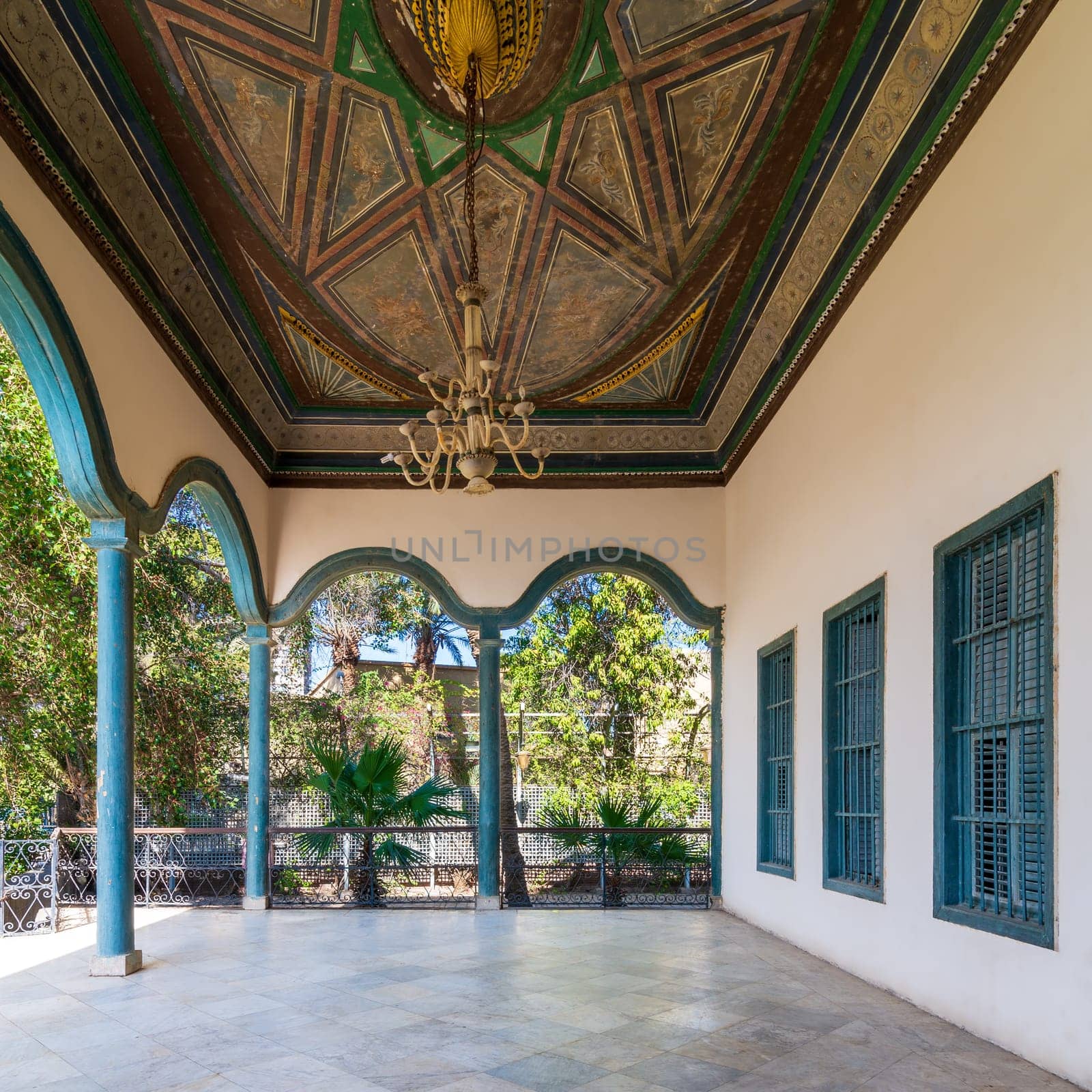 Covered terrace with decorated painted ceiling, green stone pillars, and marble floor fenced with metal forged fence and surrounded with green trees in sunny summer day