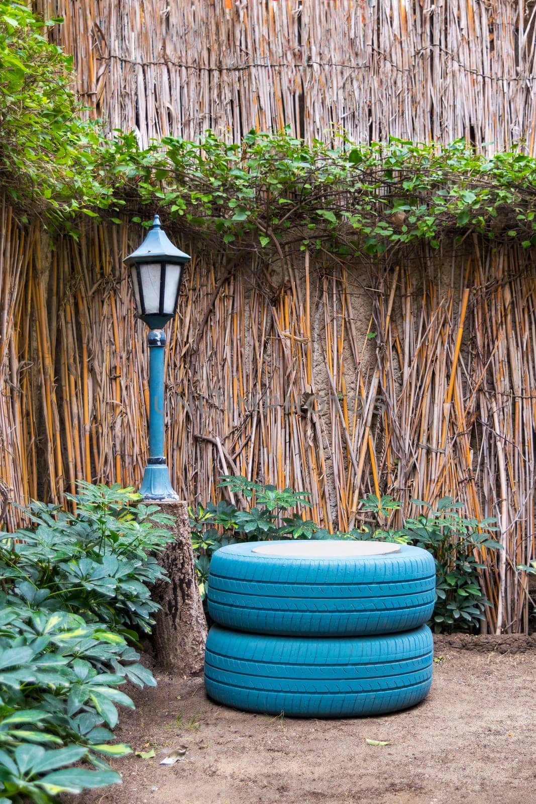 Small streetlamp and painted blue tires located near thatched fence and green plants in yard in countryside