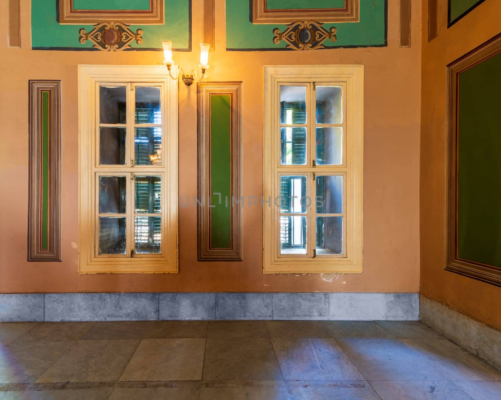 Corner orange wall with two narrow wooden window with closed green shutters, beautiful elegant rectangular green frames, and white tiled marble floor