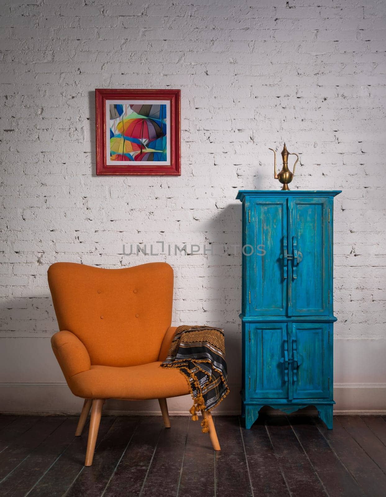 Living room corner with vintage orange armchair and ornate scarf beside blue cupboard on a red carpet, grunge dark wooden parquet floor and white bricks wall