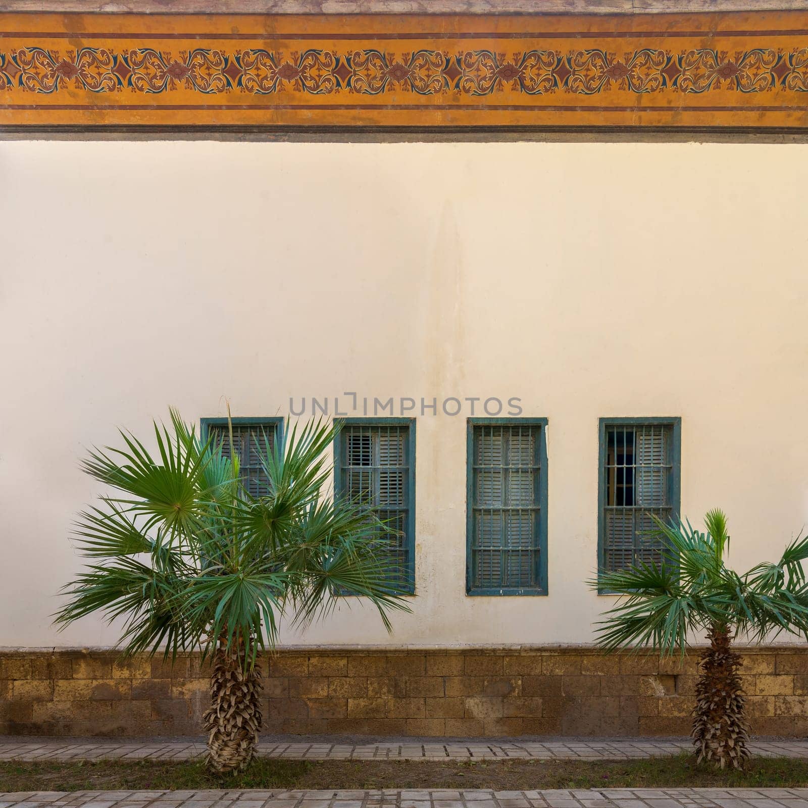 Two small palm trees and tiled stone floor in front of beige wall with orange colorful cornice decorated with floral patterns, and four green window shutters