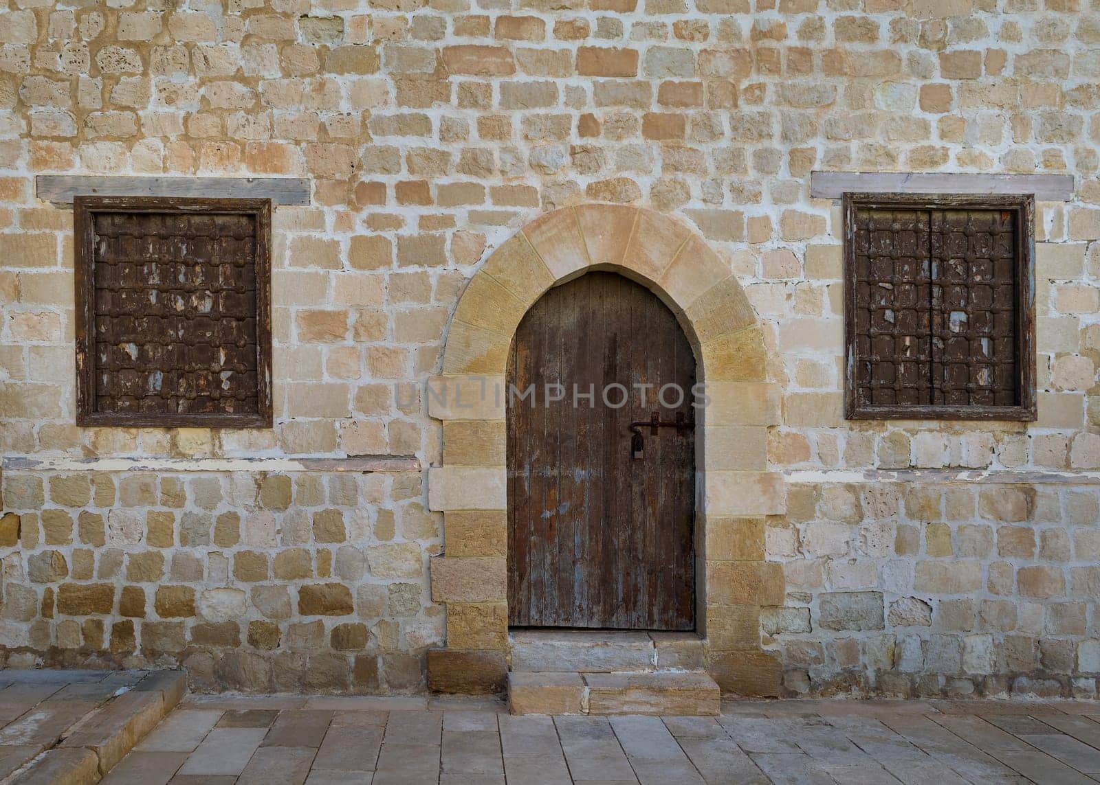 Old weathered arched wooden door and two closed rusted wrought iron windows on bricks stone wall and tiled stone floor