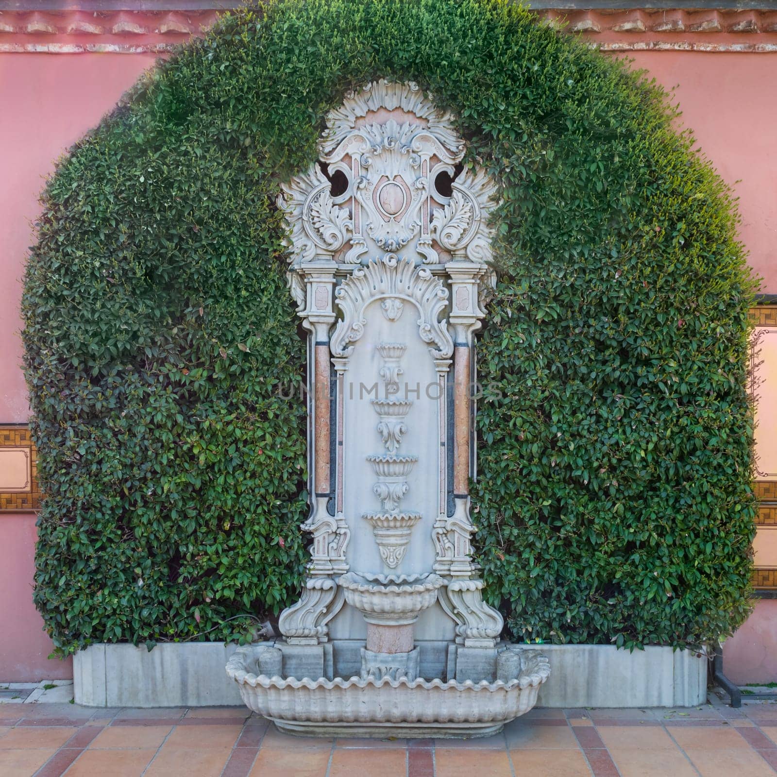 White marble sculpted wall fountain surrounded by green leaves over pink stone wall and tiled orange ceramic floor in Sultan Ahmed square, Istanbul, Turkey