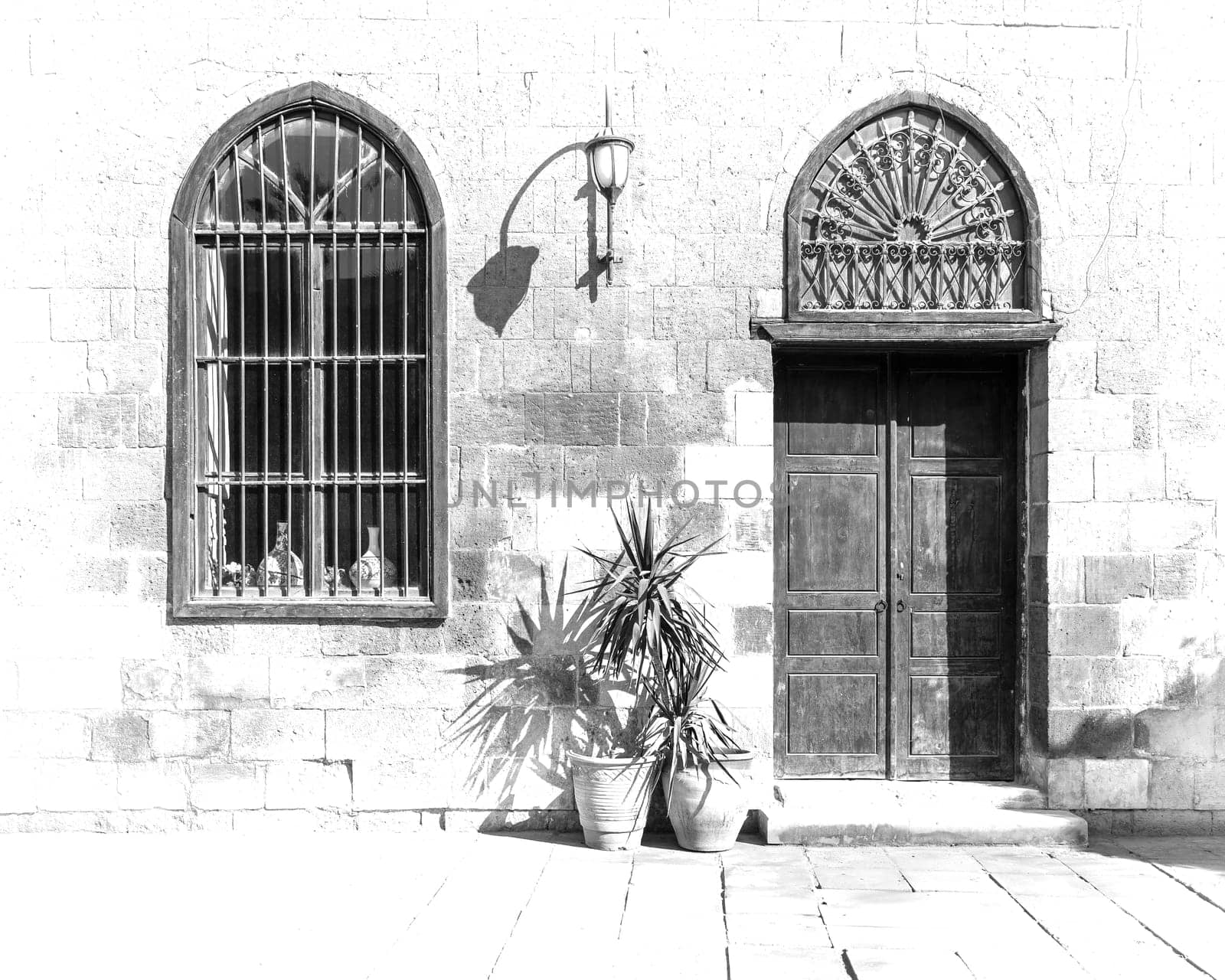 Black and white high contrast shot of old abandoned stone bricks wall with arched wooden door and window covered with wrought iron bars and lantern in a sunny day, Cairo, Egypt