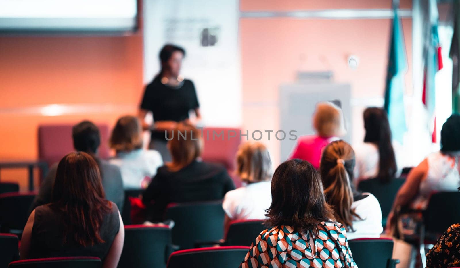 Business and entrepreneurship symposium. Female speaker giving a talk at business meeting. Audience in conference hall. Rear view of unrecognized participant in audience.