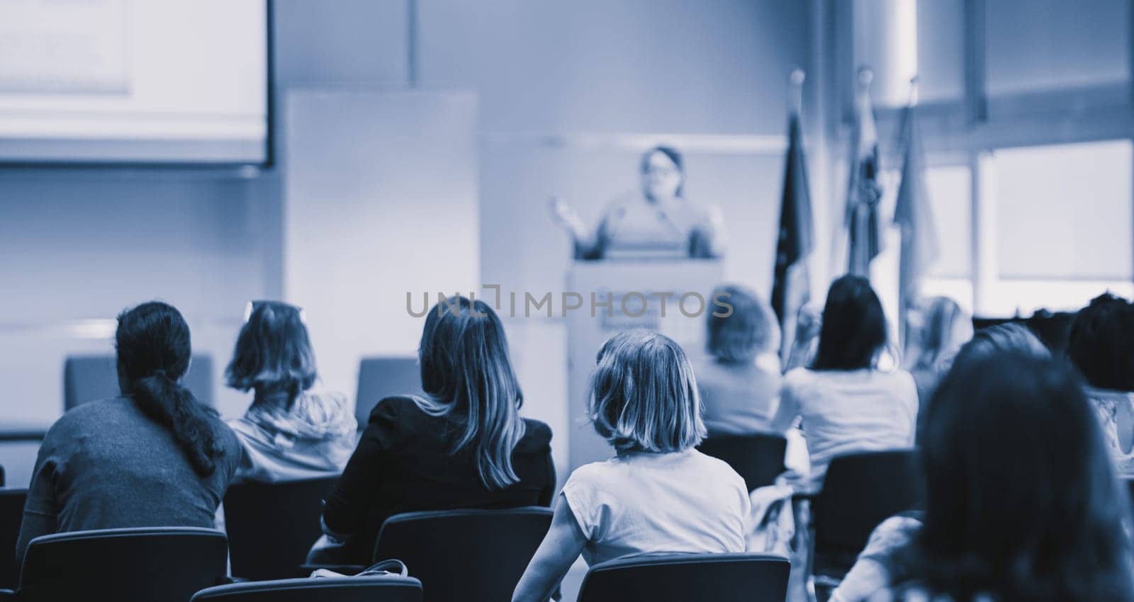Business and entrepreneurship symposium. Female speaker giving a talk at business meeting. Audience in conference hall. Rear view of unrecognized participant in audience.
