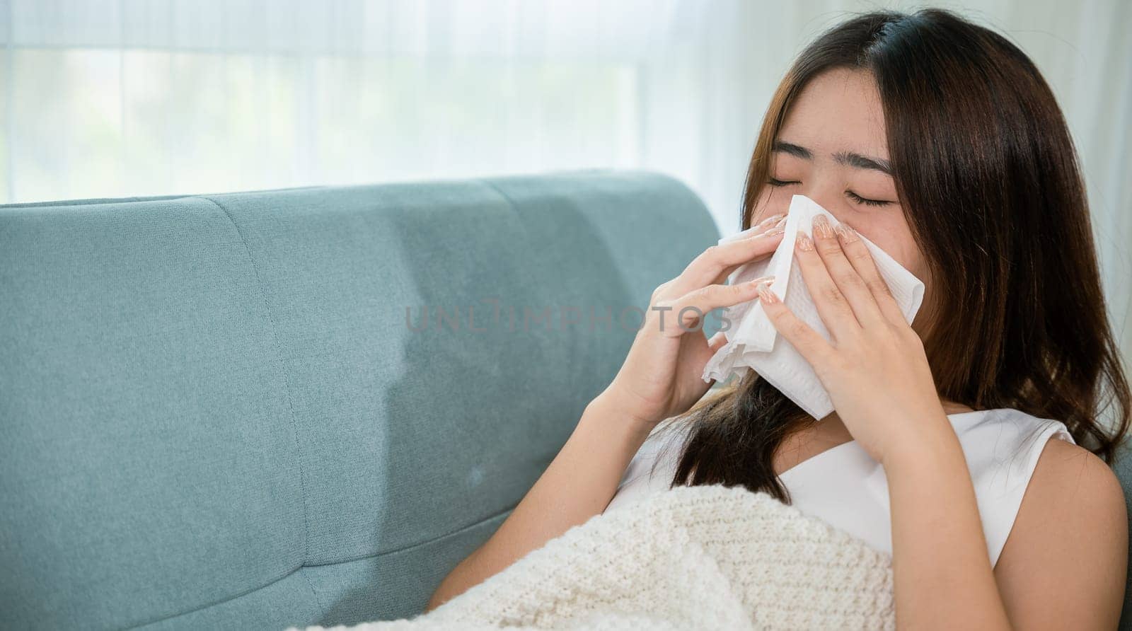Ill Asian young woman cold covered with blanket sitting on sofa and sneeze with tissue paper at home, Female sick allergic blowing nose sneezing in tissue at home, Flu health care