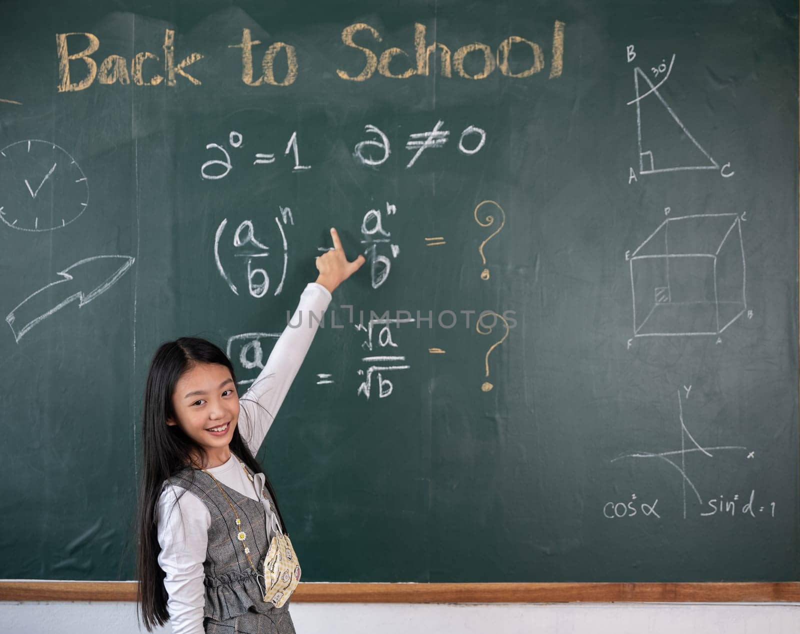 Education Back to school concept. Asian school girl in uniform with backpack on classroom pointing up finger on blackboard, primary child is standing front of class in school thinking of great ideas