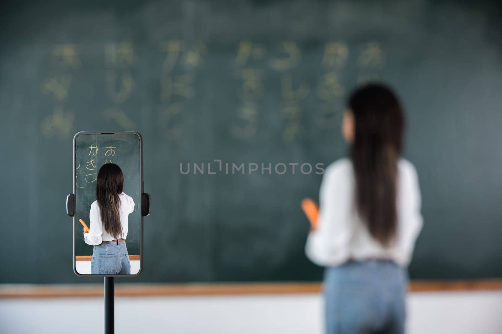 Back to school online class. Asian teacher teach on board while have blackboard lecture online through smartphone in classroom, woman standing front of class live video call online smart mobile phone