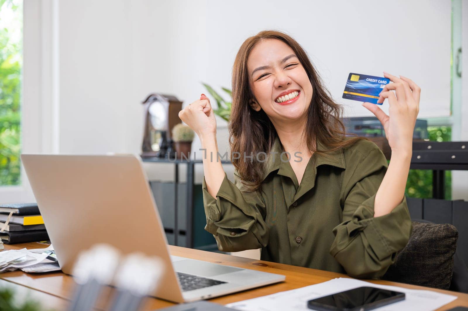 Happy Asian freelancer holding credit card she excited raised hands say yes! do winner gesture on desk home office, lifestyle business woman using laptop computer rejoicing to purchase shopping online