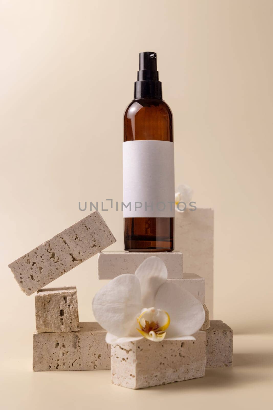 Dark glass cosmetic bottle with blank label near white orchid flowers and stones on light yellow, close up, mockup. Skincare handmade beauty product, tonic or lotion. Exotic natural cosmetics..
