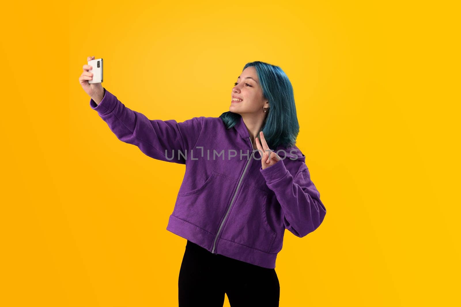 Cheerful nice young girl with blue hair taking selfie on cellphone and smiling. Isolated on yellow baclground