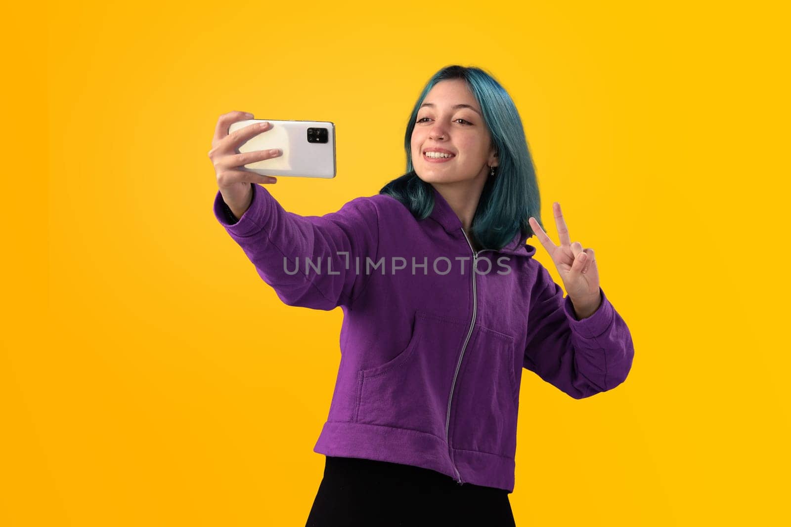 Cheerful nice young girl with blue hair taking selfie on cellphone and smiling. Isolated on yellow baclground