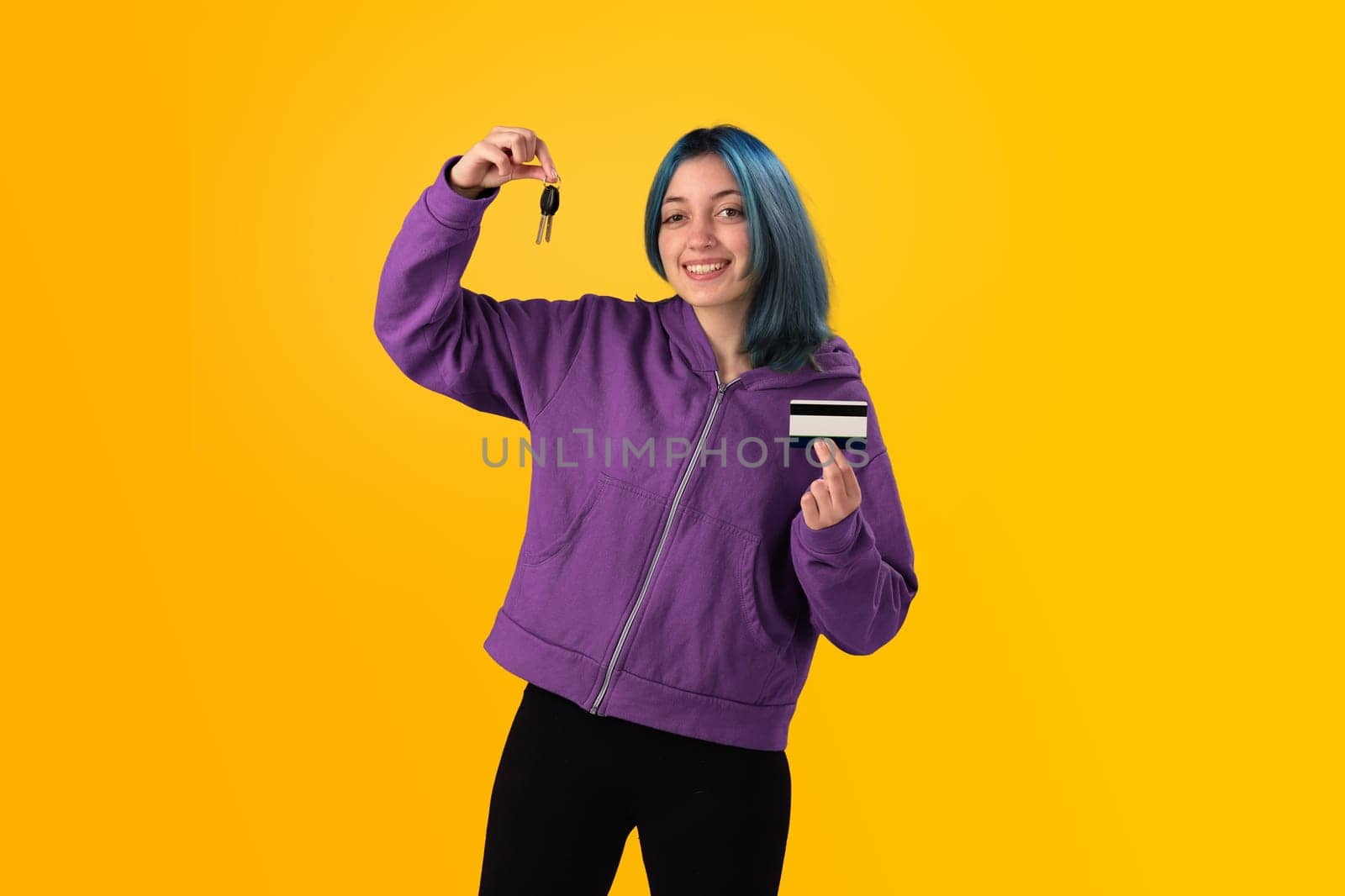 Smiling young woman student with blue hair holds a credit card and key over a yellow background