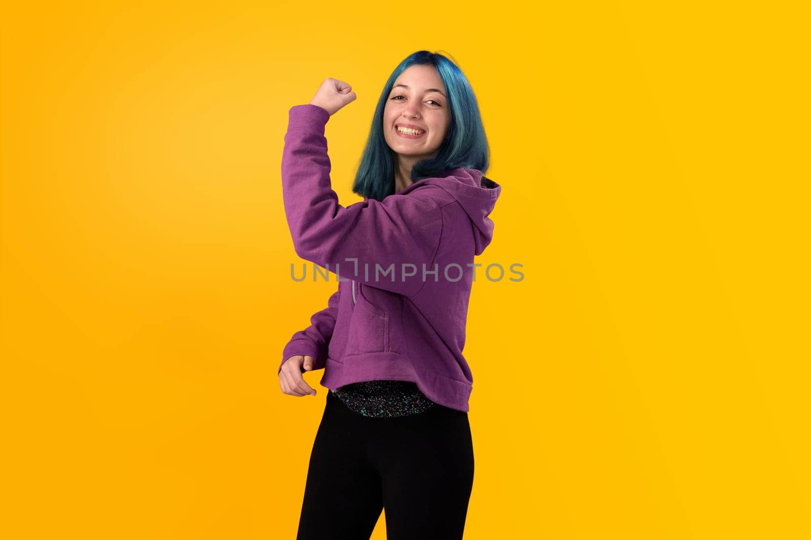 Cute young gilr with blue hair doing winner gesture, celebrate clenching fists. Isolated on plain yellow background studio portrait.
