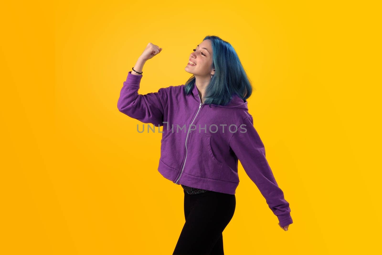 Cute young gilr with blue hair doing winner gesture, celebrate clenching fists. Isolated on plain yellow background studio portrait.