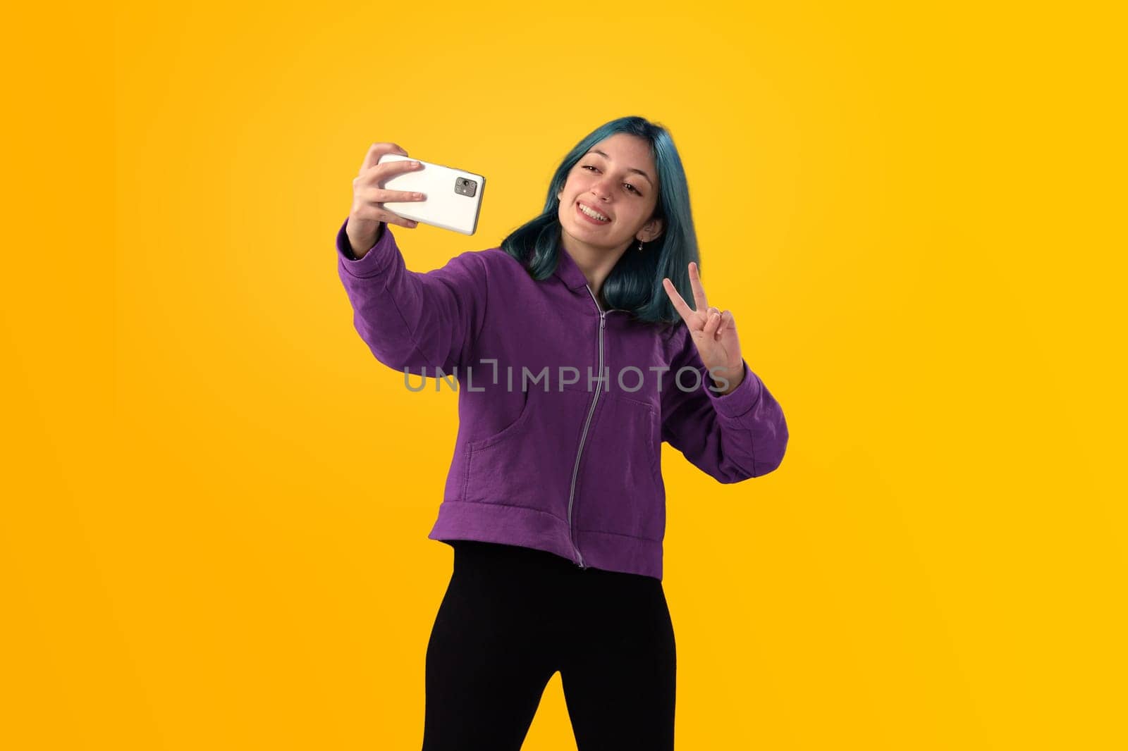 Cheerful nice young girl with blue hair taking selfie on cellphone and smiling. Isolated on yellow baclground