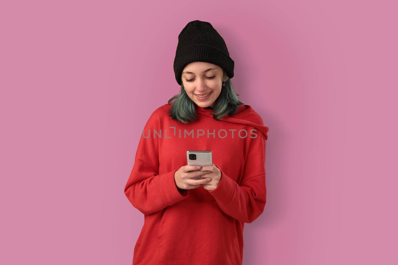 Cute young girl with blue hair using mobile phone, typing a message over pink background