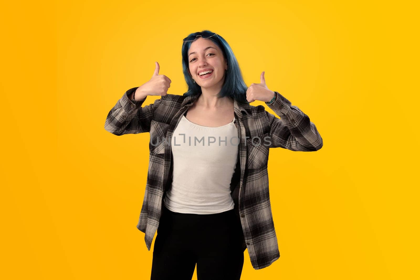 Smiling young woman student with blue hair doing positive gestures with her hands isolated over yellow background