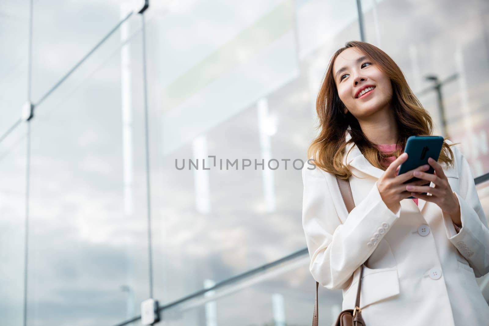 Asian businesswoman working with mobile phone standing against street front building near office, Portrait successful business woman smiling holding smartphone use application chat online in morning