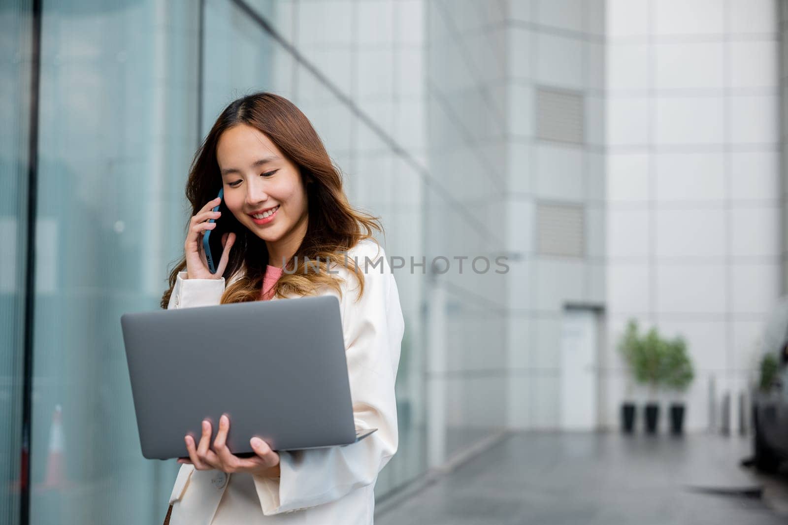 Asian businesswoman working on laptop and talking on cell phone at front building near office, Portrait beautiful business woman in white suit smile discussing issues on smart mobile phone in city