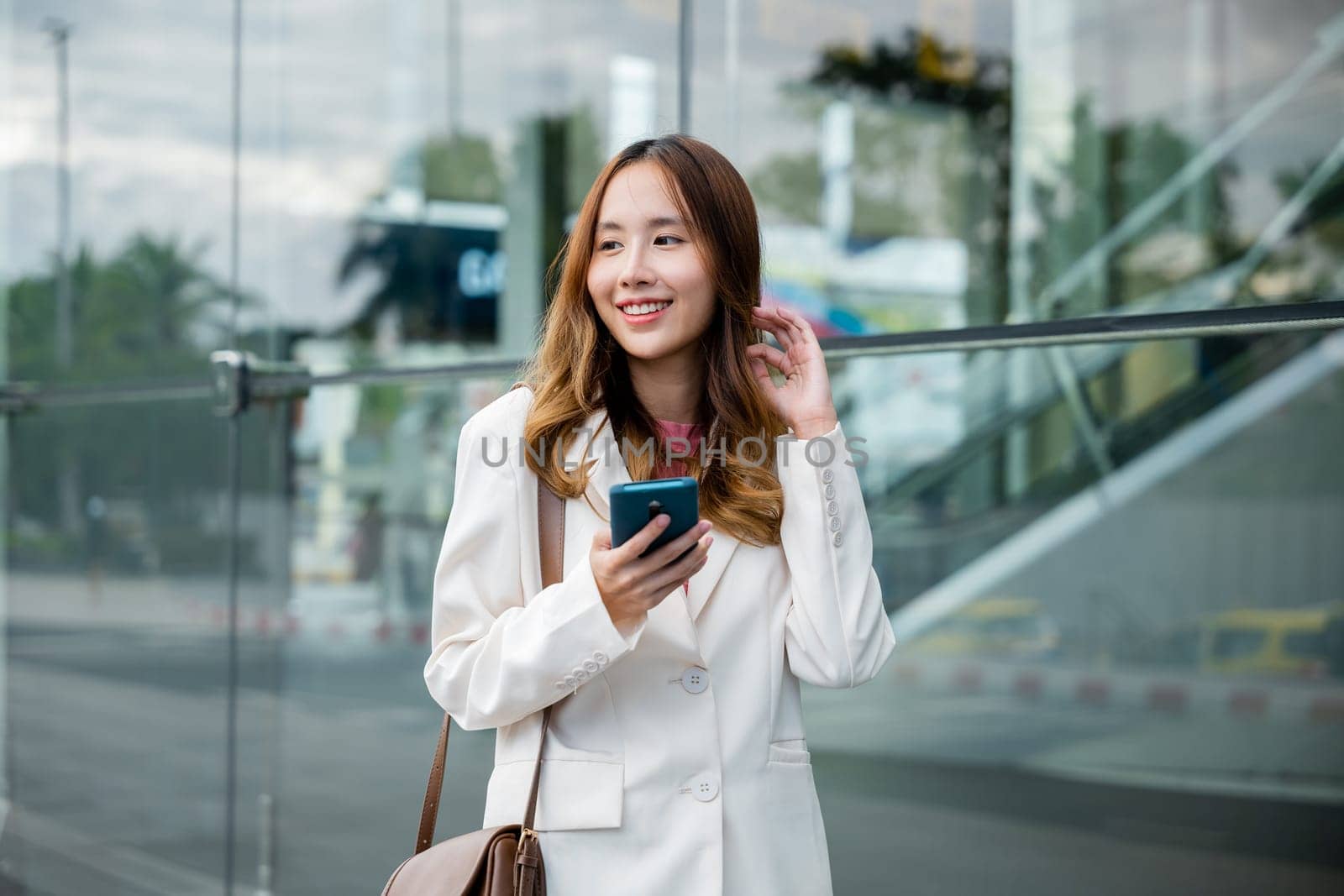 Asian businesswoman working with mobile phone standing against street front building near office, Portrait successful business woman smiling holding smartphone use application chat online in morning