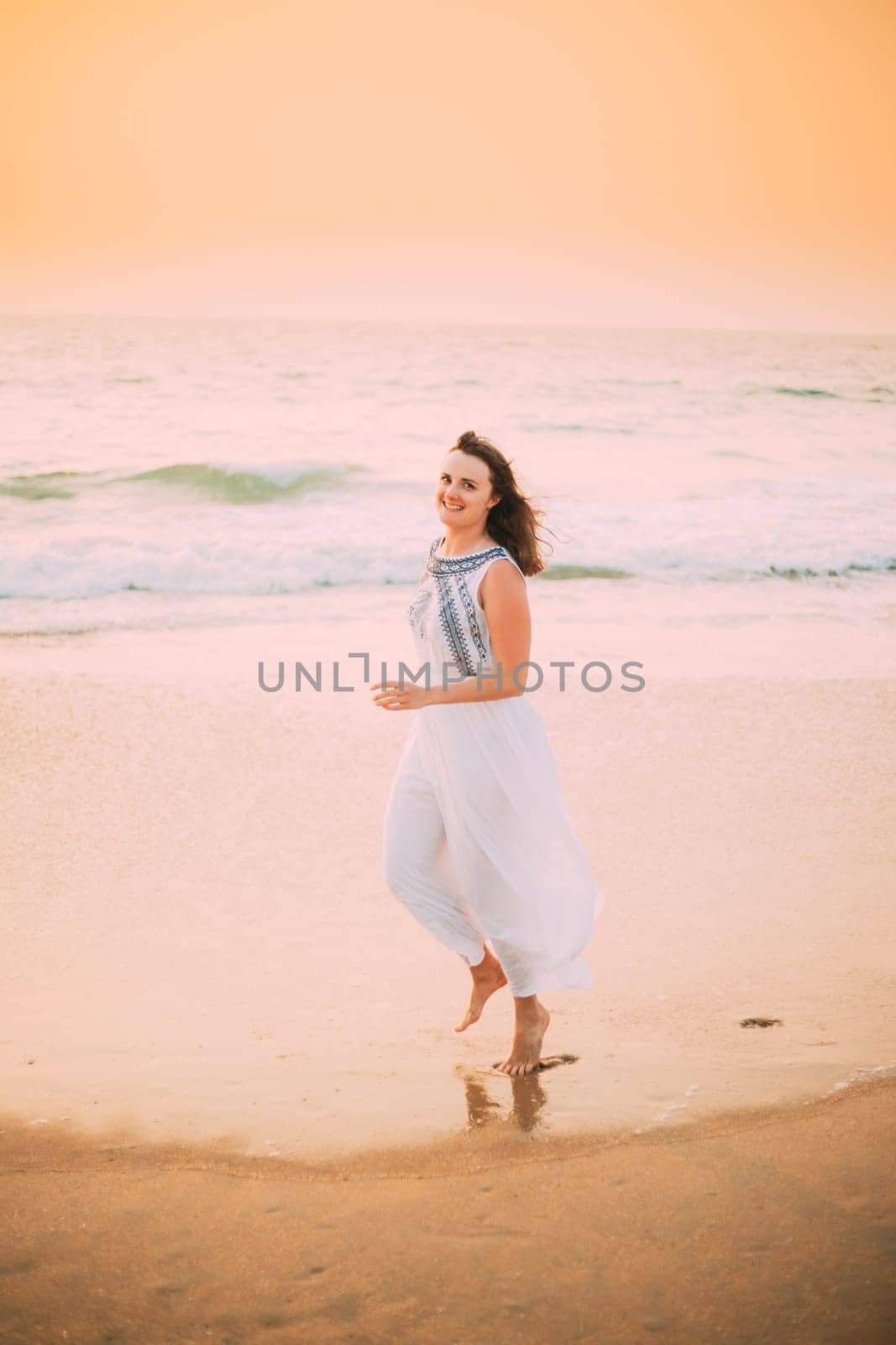 Goa, India. Young Caucasian Woman In White Dress Walking Along Seashore, Looking At Camera And Smiling In Summer Sunlight.