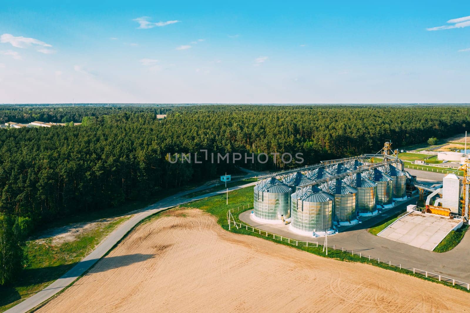 Aerial View Modern Granary, Grain-drying Complex, Commercial Grain Or Seed Silos In Sunny Spring Rural Landscape. Corn Dryer Silos, Inland Grain Terminal, Grain Elevators Standing In A Field.