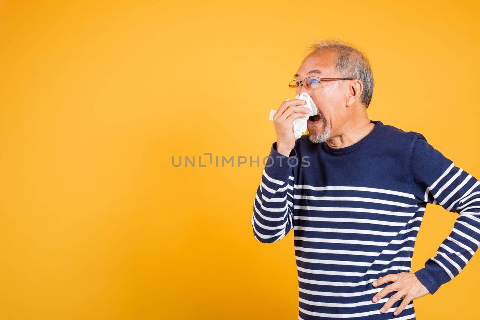 Asian elder man cold having flu and sneezing from sickness virus problem use tissue studio shot isolated on yellow background, Portrait senior old man blowing nose with tissue, pensioner unwell