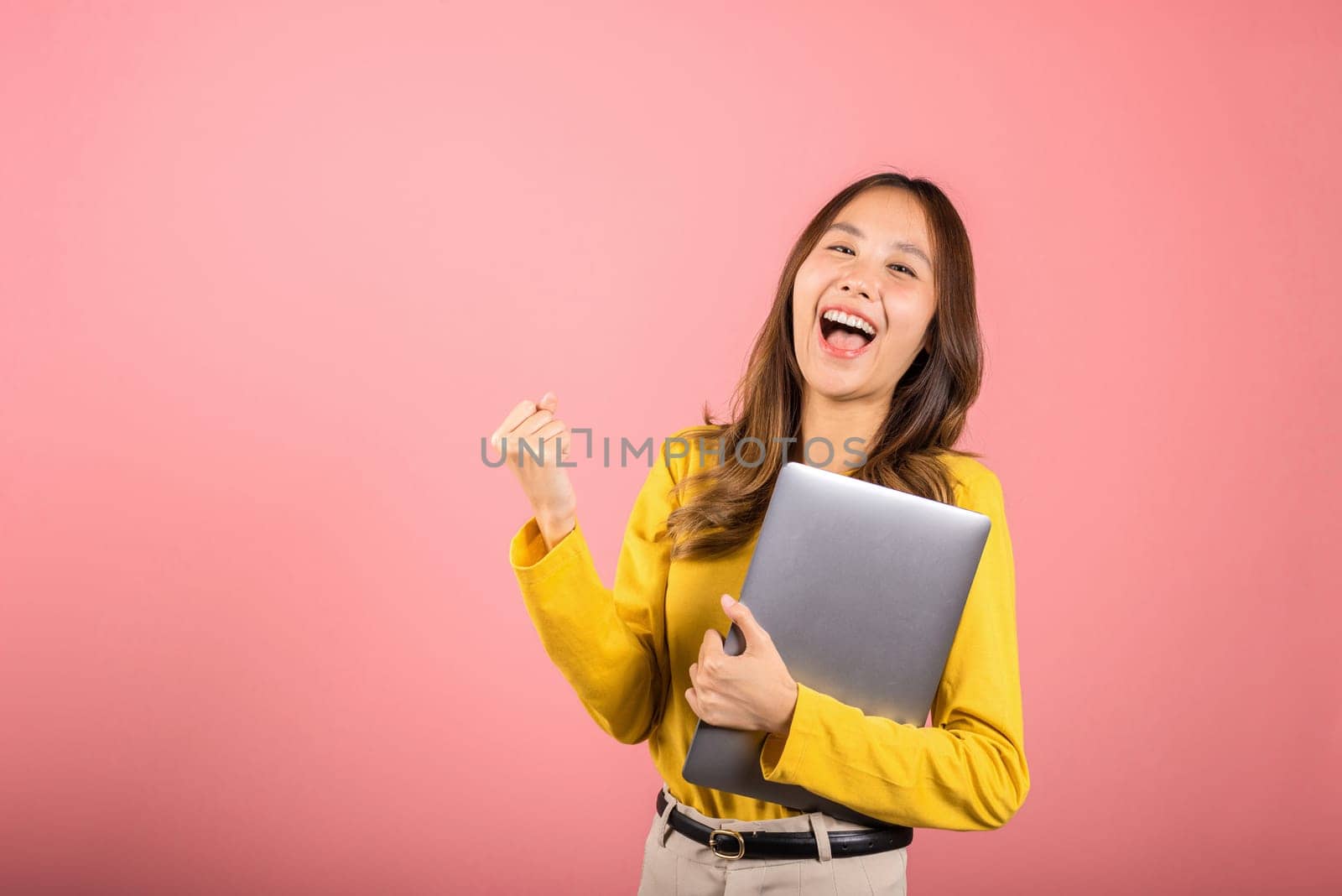 Portrait of happy Asian beautiful young woman confident smiling face holding laptop computer raise hand and say yes! excited celebrating success, studio shot isolated on pink background, copy space