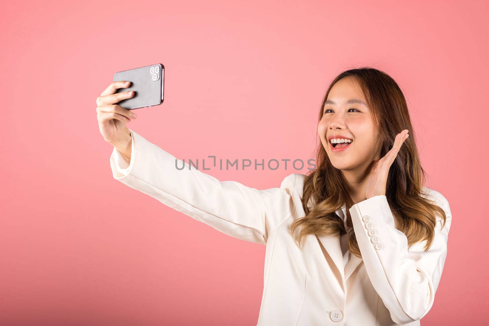 Happy Asian portrait beautiful cute young woman smiling excited  making selfie photo, video call on smartphone studio shot isolated on pink background, female hold mobile phone raise hand say hello
