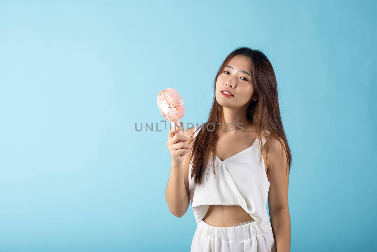 Asian beauty young woman holding pink portable electric mini fan near her face studio shot isolated on blue background, Female hand hold small plastic fan handheld she enjoying cool wind blowing