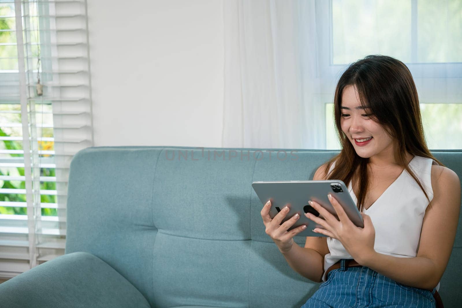 Happy Asian woman playing smart tablet at home while relaxing on sofa living room, Beautiful female sitting on couch using digital tablet pc, technology social media