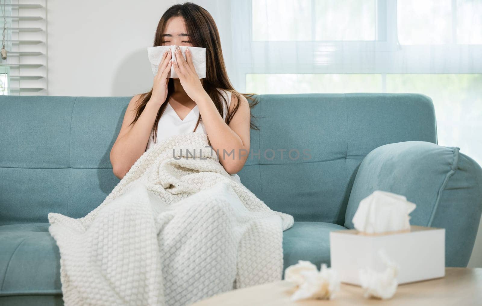 Ill Asian young woman cold covered with blanket sitting on sofa and sneeze with tissue paper at home, Female sick allergic blowing nose sneezing in tissue at home, Flu health care