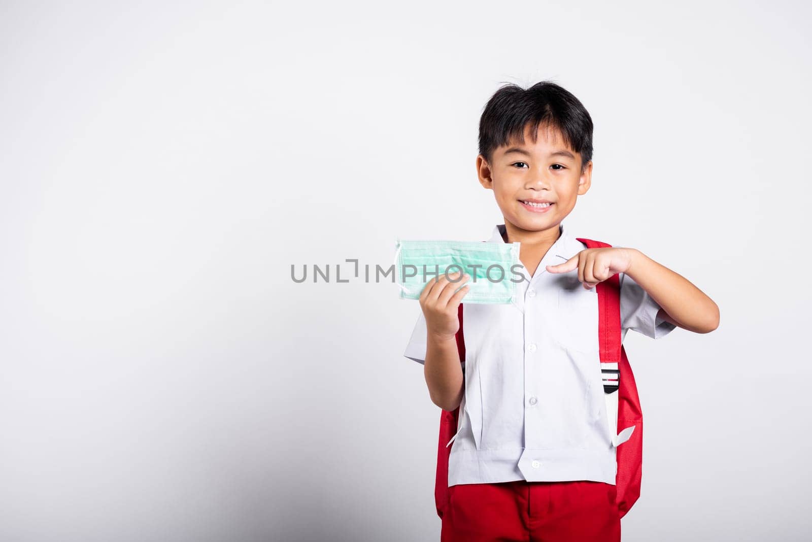 Asian student kid boy wearing student thai uniform and pointing finger to protect mask before to go to school in studio shot isolated on white background, preschool, new normal back to school