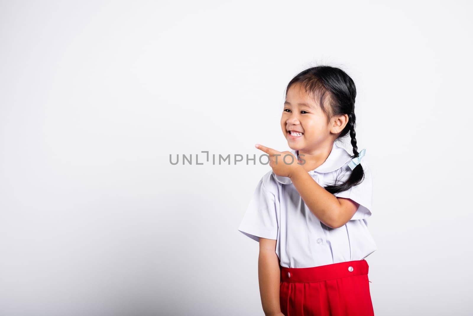 Asian toddler smiling happy wear student thai uniform red skirt keeps pointing finger at copy space in studio shot isolated on white background, Portrait little children girl preschool, Back to school