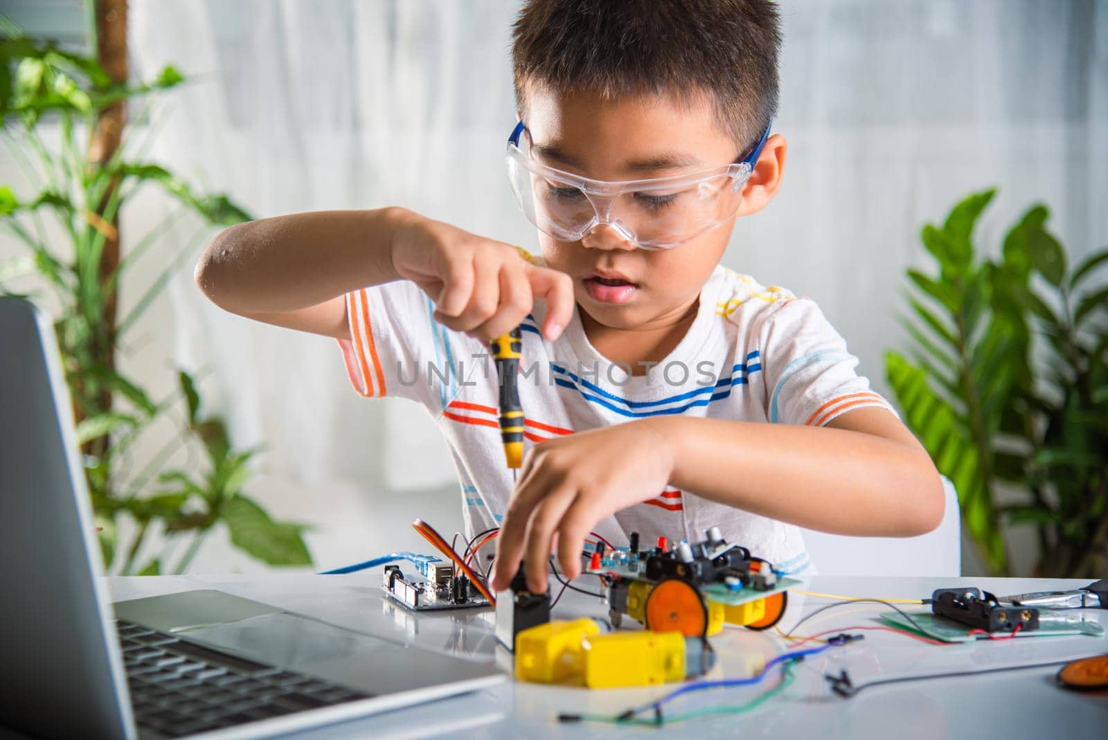 Asian kid boy assembling the Arduino robot car homework project at home, Little child tighten the nut with a screwdriver to assemble car toy, creating electronic AI technology workshop school lesson