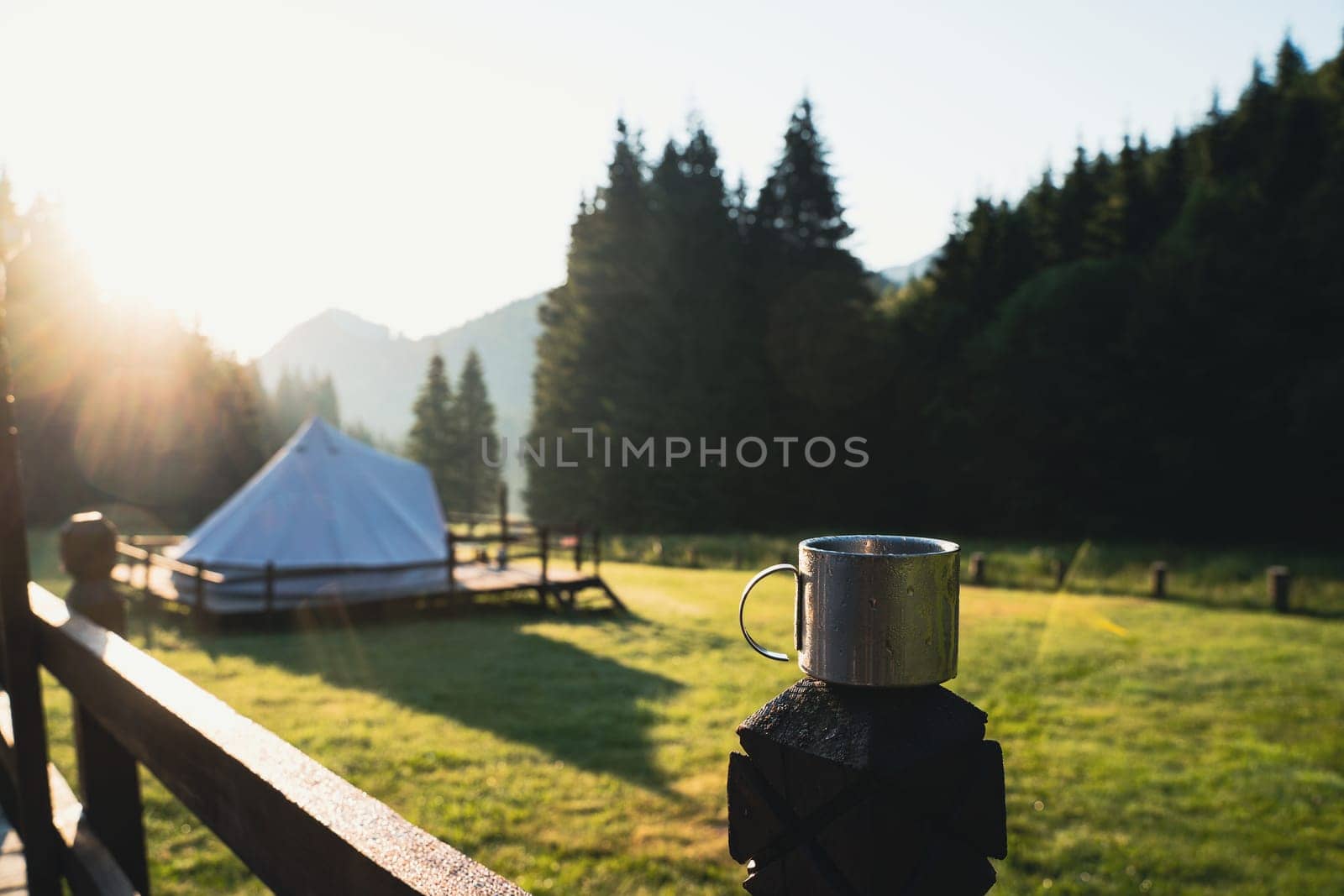 closeup front view of metallic coffee cup in the morning with glamping tent camping ground in the background in a green mountain meadow surrounded by fir tree forest