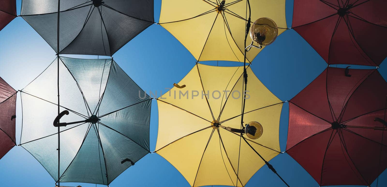 front view closeup detail of colorful umbrellas and Edison light bulbs hanging out above a city street against a blue sky