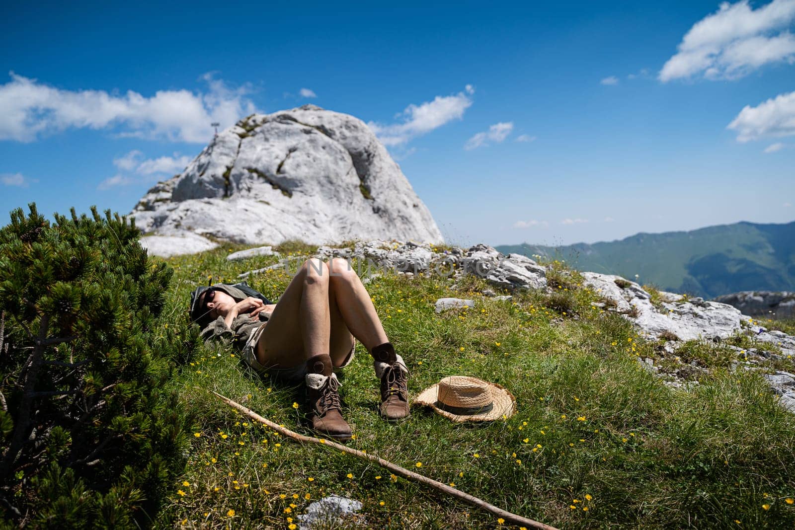 young Caucasian woman lying down and taking a rest in the warm sun near a wooden trekking stick and wicker hat in the grass on a mountain peak with blue sky and clouds above