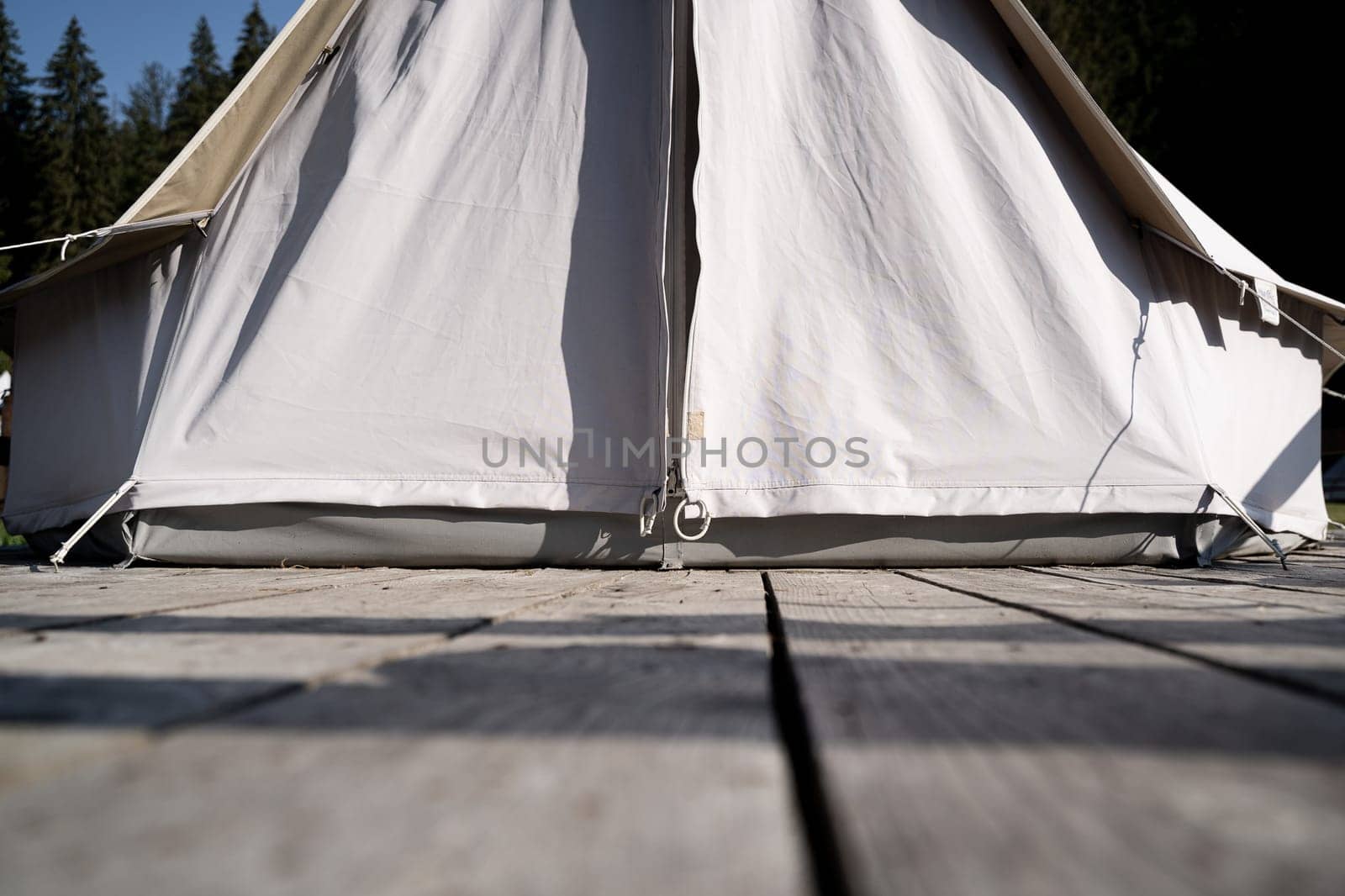 font view closeup of glamping tent white fabric entrance on wooden deck in green meadow surrounded by fir tree forest