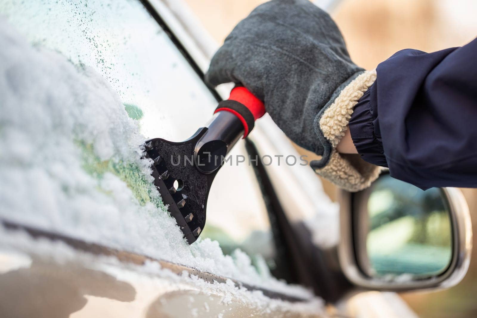 Hand in glove holding a snow scraper and removing ice from a car window with rear view mirror in background. Concept of danger on roads in winter season. Maintenance of vehicle in cold temperatures.