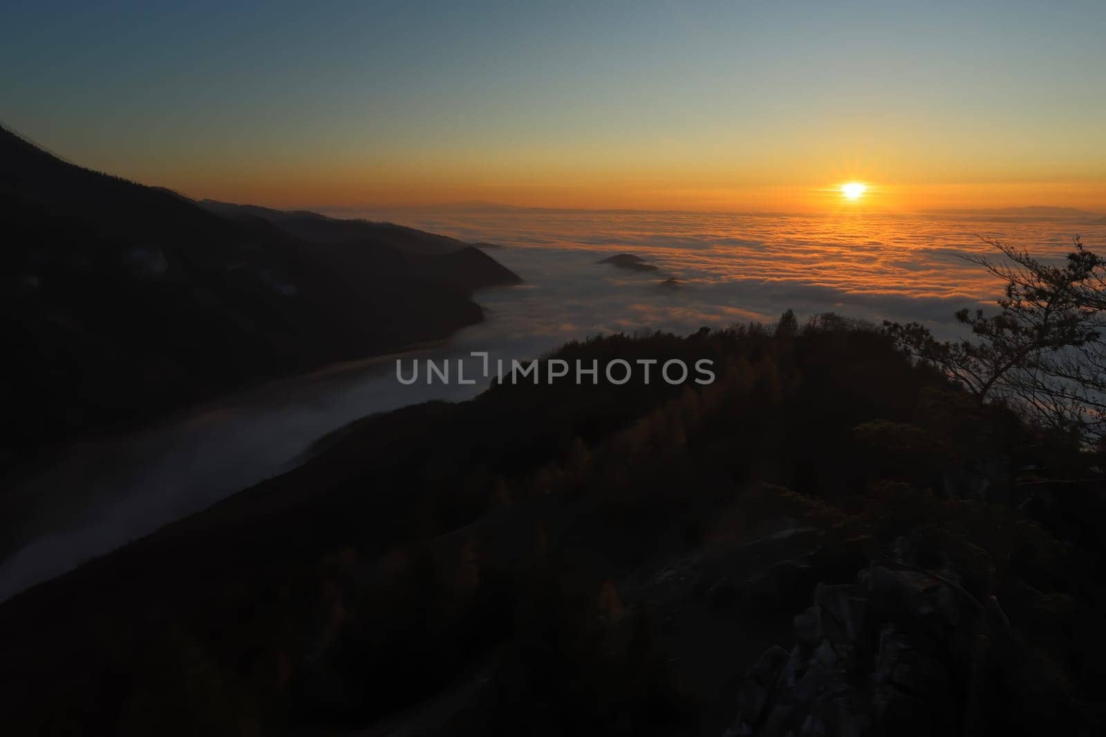 Sun rising above low clouds in mountain with dark hills in background. Nature scenery early in the morning with illuminated sky and valley still hidden in shadow.