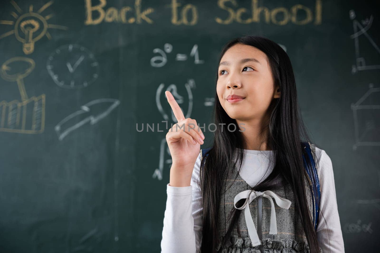 Education Back to school concept. Asian school girl in uniform with backpack on classroom pointing up finger on blackboard, primary child is standing front of class in school thinking of great ideas