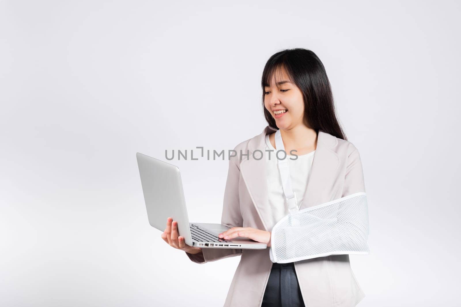 Asian business woman confident smiling broken arm after accident and wear arm splint for treatment but still have to work she hold laptop computer studio isolated on white background,copy space