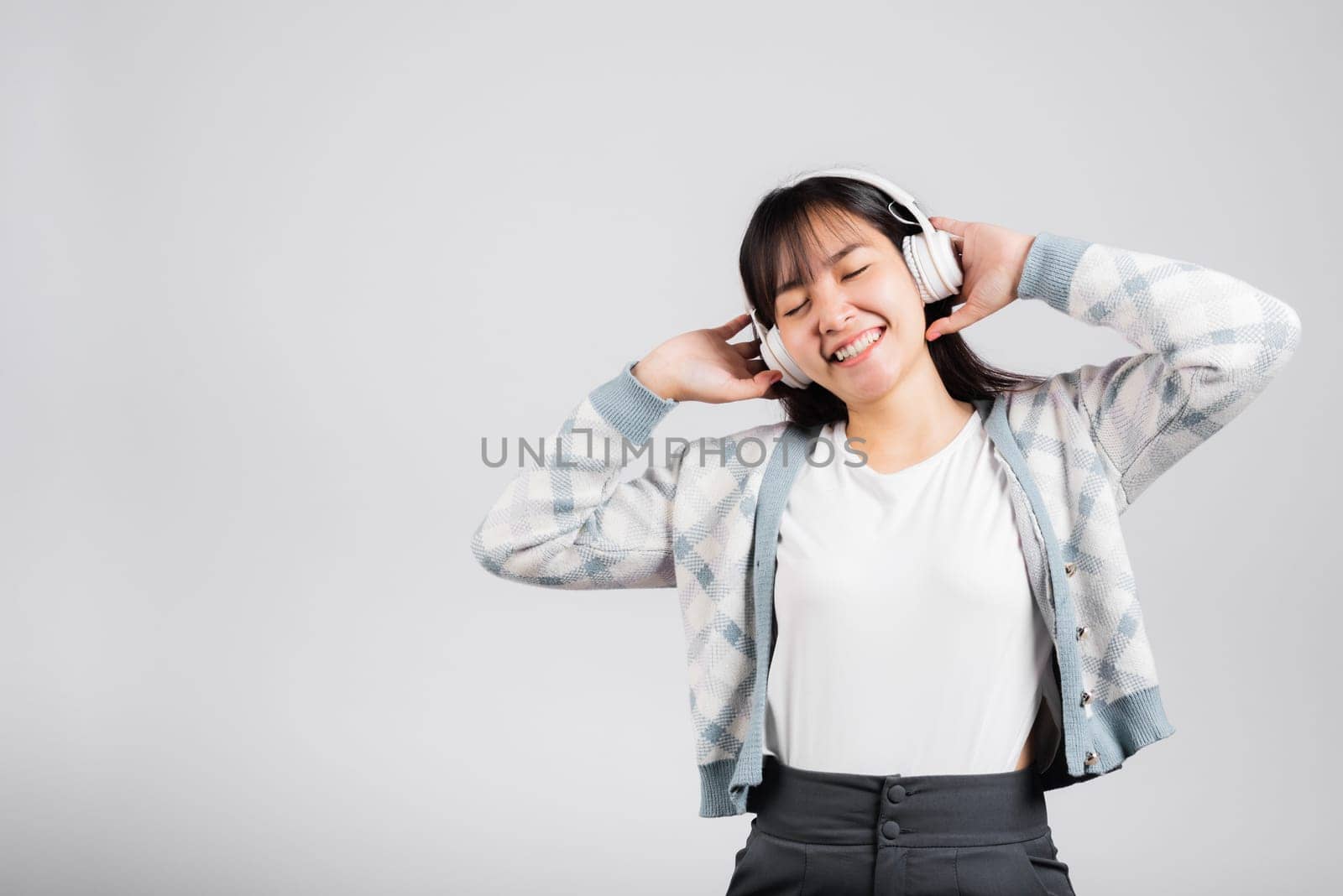 Woman excited smiling listening to music radio in bluetooth headphones and holding phone studio shot isolated white background, happy Asian young female listen modern technology mobile phone